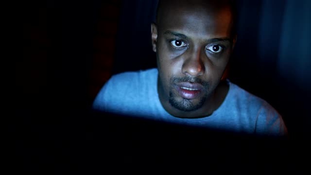 一个非洲人晚上在黑暗中上网。看着笔记本电脑工作到很晚的黑人视频素材