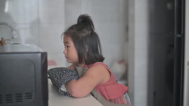 年轻的亚洲中国女孩在厨房里戴着防护手套等着，从微波炉里看她的食物视频下载