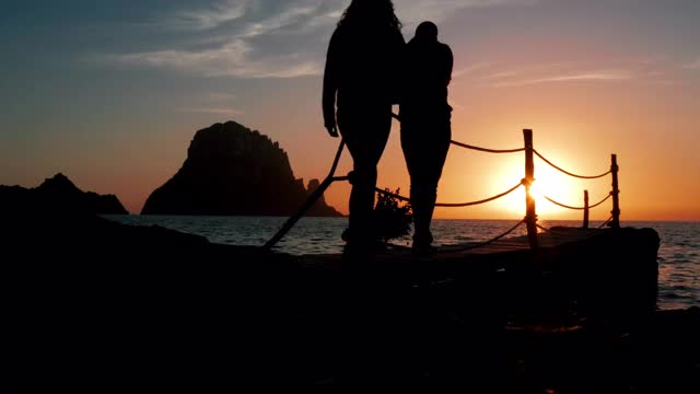 在夕阳下，一对年轻夫妇手牵手穿过一个木制码头，并与背景中的“Es Vedrà”接吻——4K视频下载