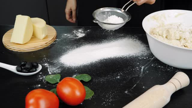 在专业的现代厨房里，女人正在往黑色的表面上倒白面。面粉正从黑桌上的筛子中筛出来。准备披萨面团视频素材