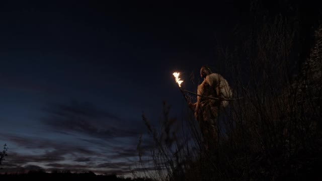 原始的狩猎采集者，穿着羊皮，手持弓箭，晚上在农村用燃烧的木棍点燃火把行走。原始穴居人的肖像穿着动物夜视频素材