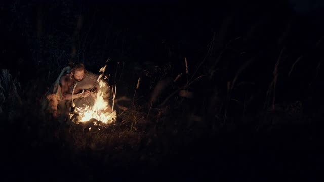 男性野蛮人坐在附近燃烧的火和磨刀在晚上在野外露营视频素材