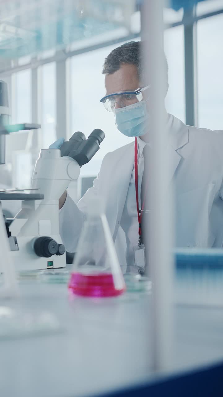 垂直视频。医学发展实验室:科学家戴着口罩在显微镜下观察，输入数据到数字平板电脑。高级实验室医学、生物技术研究视频素材