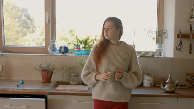 一个年轻女人拿着茶杯站在厨房里视频素材