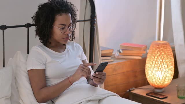 成功，非洲妇女在床上用智能手机庆祝视频素材