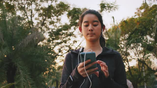 迷人的亚洲少女听音乐，同时在公园里跑步。年轻女子晨跑锻炼。视频下载
