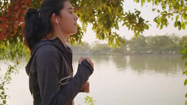 中镜头:迷人的亚洲少女听音乐，同时在公园的湖上奔跑。年轻女子晨跑锻炼。视频素材