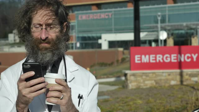 留胡子的男医生在医院外阅读和使用他的智能手机视频下载