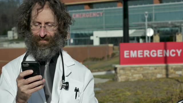 留着胡子的英俊男医生在医院外使用手机视频下载