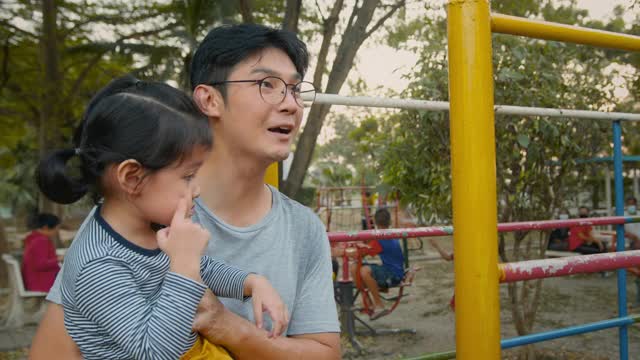 亚洲父亲和他的女儿在公园的操场上玩。学步的小女孩在周末和父母一起玩耍，享受和感受快乐。家庭在公园的概念视频购买
