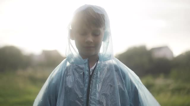 可爱自信的男孩拉着雨衣，看着相机站在雨天的户外。可爱的白人孩子的肖像在日落在阳光户外摆姿势。快乐的童年概念视频下载