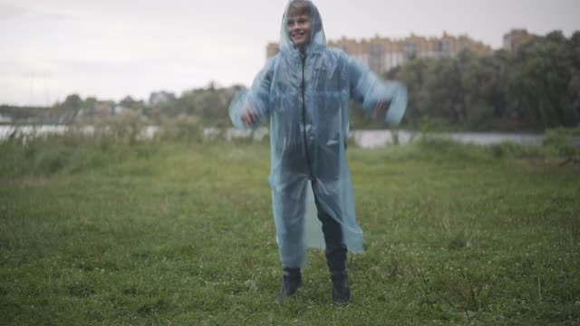 照相机接近快乐的白人男孩在雨衣有乐趣在雨天的夏天户外。活泼可爱的男生的肖像跳跃和旋转。快乐的童年概念视频下载