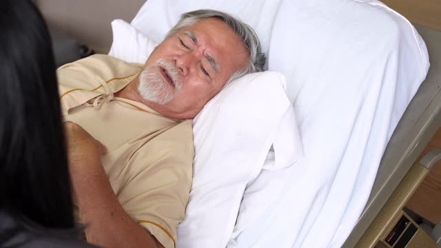 亚洲老年男性病人躺在现代医院康复室的床上，与女儿谈论心脏病的症状。医院医疗中心和家庭医疗保险概念。视频素材