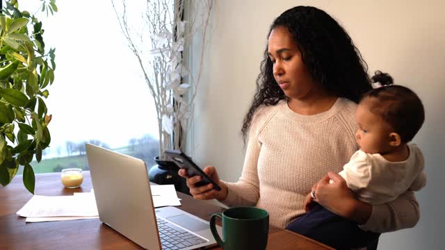非裔美国母亲抱着她的混血婴儿女儿，一边用手机发短信一边用手提电脑，马克杯和文件坐在家里餐桌桌上临时工作的桌子上。视频素材