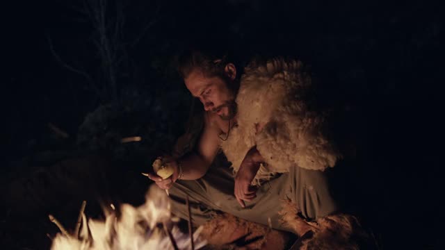 穿着兽皮的人用石头磨刀，然后在晚上坐在篝火旁边小心地环顾四周。男性野蛮人在火边磨刀视频素材
