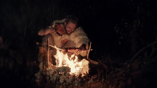 一个披着兽皮的大胡子男人，晚上在乡下，在火边休息，吃苹果。晚上在火边吃东西的野蛮人视频素材
