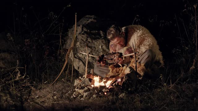 饥饿的野蛮人在篝火旁吃肉。饥饿的人贪婪地吃着炸肉，而休息在篝火在黑暗的夜晚在自然界视频素材