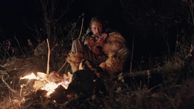 饥饿的野人在篝火旁吃东西。低角度饥饿的男性野蛮人吃肉，而坐在附近的火在晚上在农村视频素材
