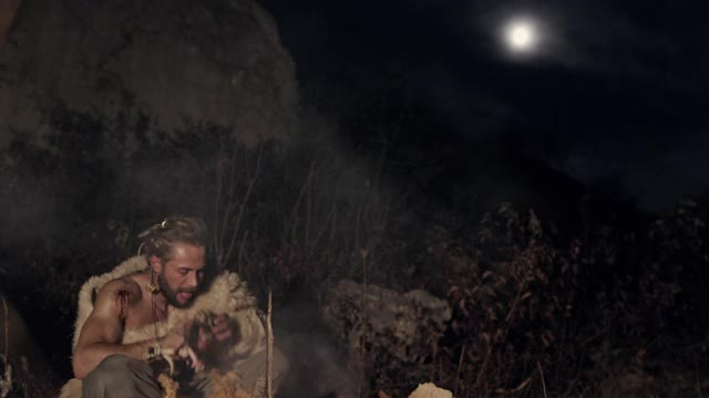 饥饿的穴居人尼安德特人或克鲁马努野蛮人猎人吃油炸肉和看着月亮，休息在篝火附近晚上视频素材