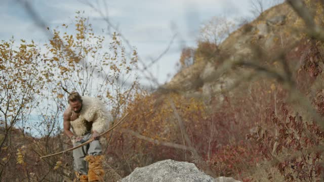低角度的野蛮弓箭手在兽皮跳上石头然后瞄准弓和射箭在阴天的自然狩猎视频下载