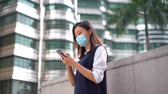 年轻的亚洲女性戴着防护口罩，使用智能手机视频下载