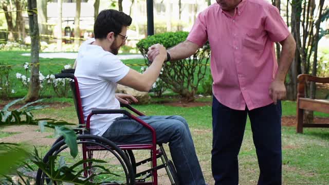 爸爸在公园里帮助坐在轮椅上的儿子视频下载