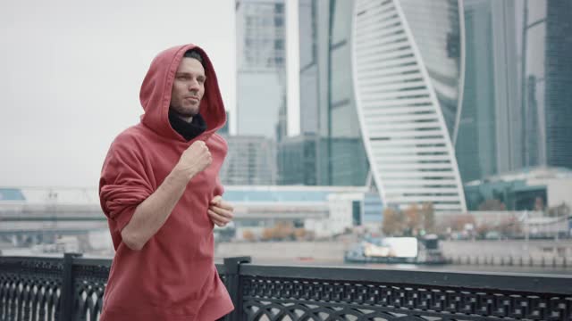 穿着红色连帽衫的商业摩天大楼背景上的跑步者视频下载