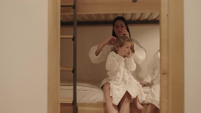 妈妈和两个穿浴袍的孩子坐在双层床上，把女孩的头发扎成马尾辫视频下载