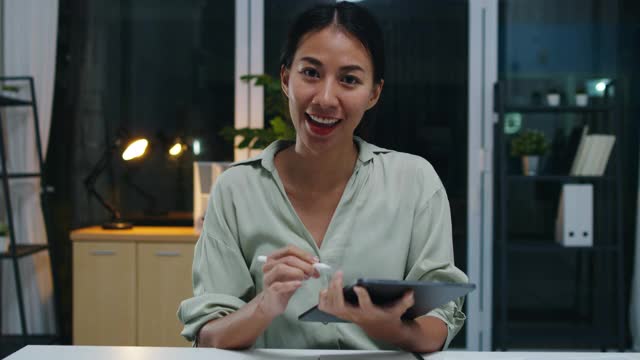 亚洲商界女性社交距离成为病毒预防的新常态，在晚上工作的时候看着摄像机向同事展示视频通话计划。视频素材