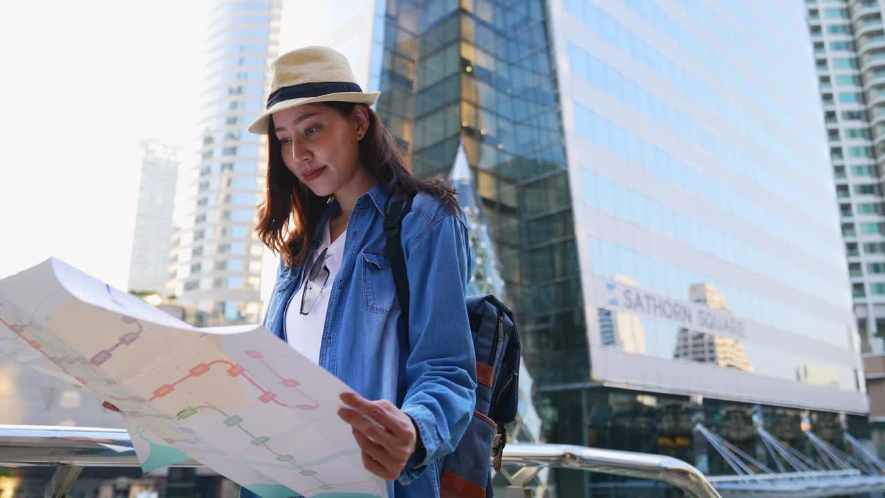 一位穿着旅游服装的美丽亚洲女子正在摊开一张以大城市为背景的地图。视频下载