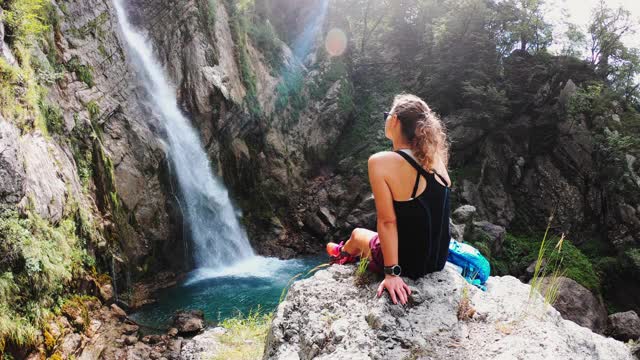 一个女人坐在一个美丽的瀑布旁边的岩石上的后视图视频下载