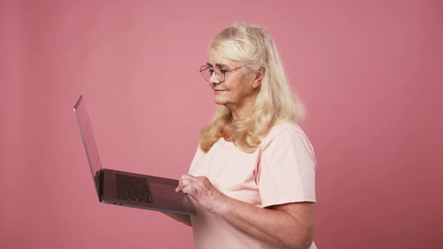 快乐兴奋的高级妇女戴着眼镜在网上工作笔记本电脑，享受成功，粉红色的工作室背景视频素材