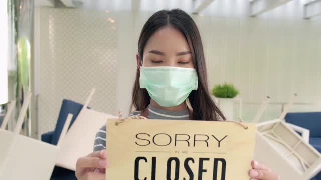 年轻的亚洲女服务员穿着围裙，戴着安全面具走在咖啡馆里，同事在用布擦桌子作为背景。中小企业在检疫期间的概念。视频素材