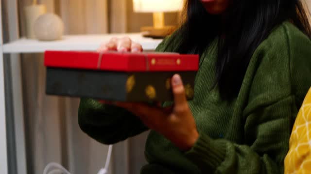 快乐的亚洲女孩穿着绿色毛衣和圣诞老人帽坐在沙发上打开圣诞礼盒在家里，节日的概念。视频下载