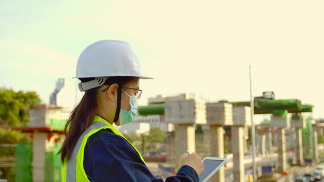 亚洲女性建筑建筑师或土木工程师专业人员戴着安全安全帽在城市建设现场的房地产建筑项目中工作，在平板上检查和记录建设进度。视频素材