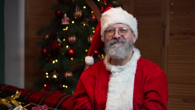 圣诞老人看着镜头，竖起大拇指。在挂着玩具和彩灯的圣诞树的背景下，穿着红色衣服和帽子的胡子老人。慢动作视频下载