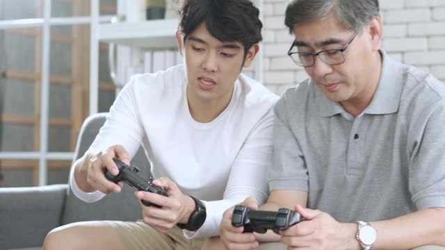 年长的亚洲男人在退休年龄有乐趣玩视频游戏机与他的儿子在他的舒适的家视频下载
