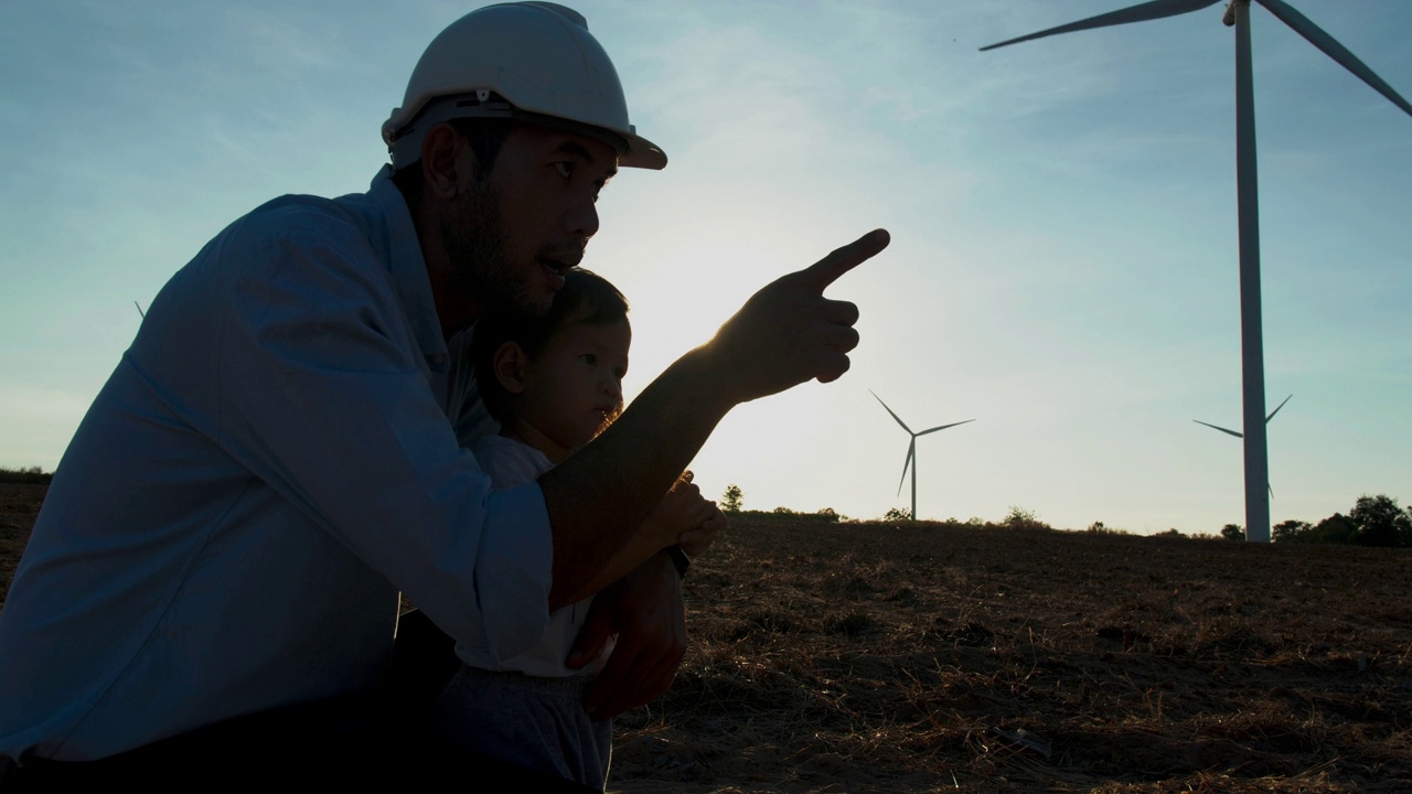 亚洲父亲剪影日落是在风力发电站工作的工程师，和15个月大的男婴一起看着风力发电机的背景。教育孩子关于可持续发展的概念。视频下载