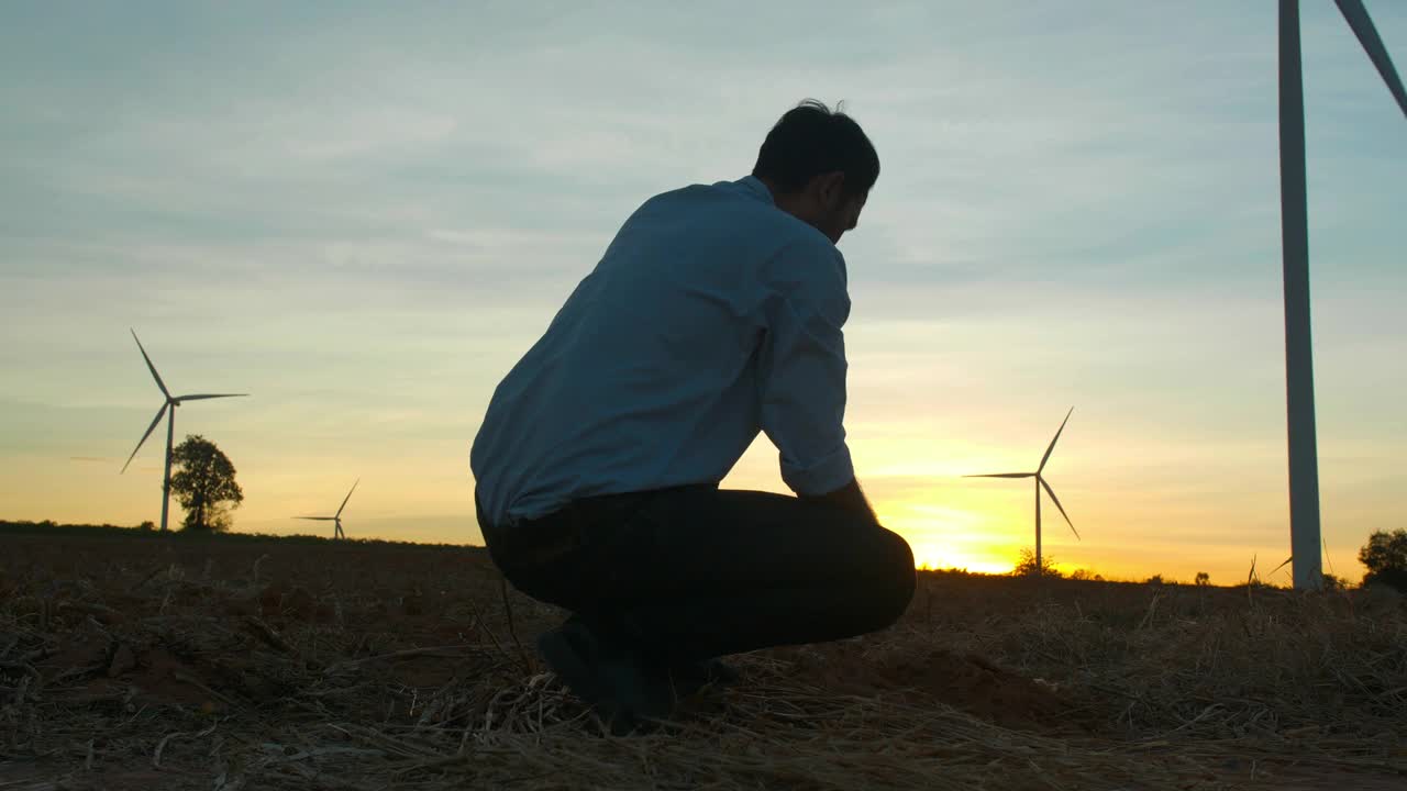 在风力发电站工作时，年轻的男工程师在检查风向和土壤的剪影日落，检查风力农场的能源生产。可再生能源工厂的概念。视频素材