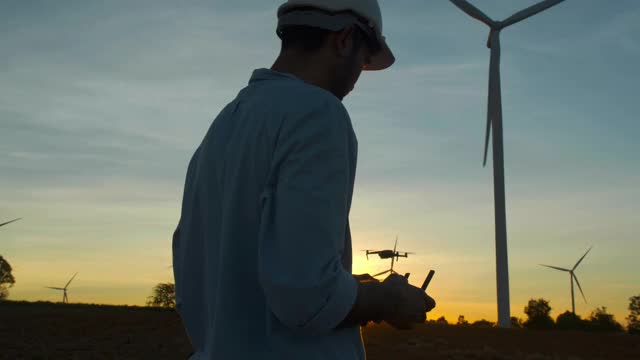 日落的剪影，年轻的男性工程师控制无人机在风力发电站工作，检查风力发电厂的能源生产。可再生能源工厂的概念。视频素材