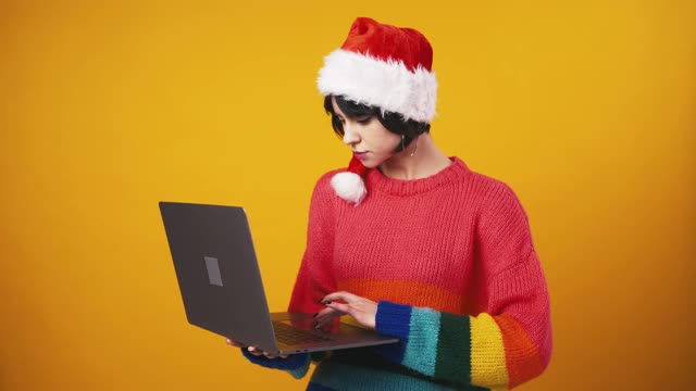 可爱的女士戴着圣诞老人的帽子在笔记本电脑上浏览，享受在线的胜利，发现伟大的应用程序，橙色的工作室背景视频素材