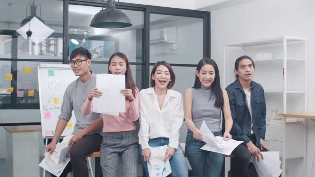一群亚洲年轻创意人士穿着时尚休闲装庆祝项目成功并将文件扔进办公室。不同的亚洲男性和女性站在一起创业。视频素材