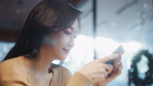 美丽年轻的亚洲女性在咖啡店使用智能手机，快乐的微笑。人们现代的生活方式，互联网的通讯技术，或网上购物的概念。手持慢动作视频下载