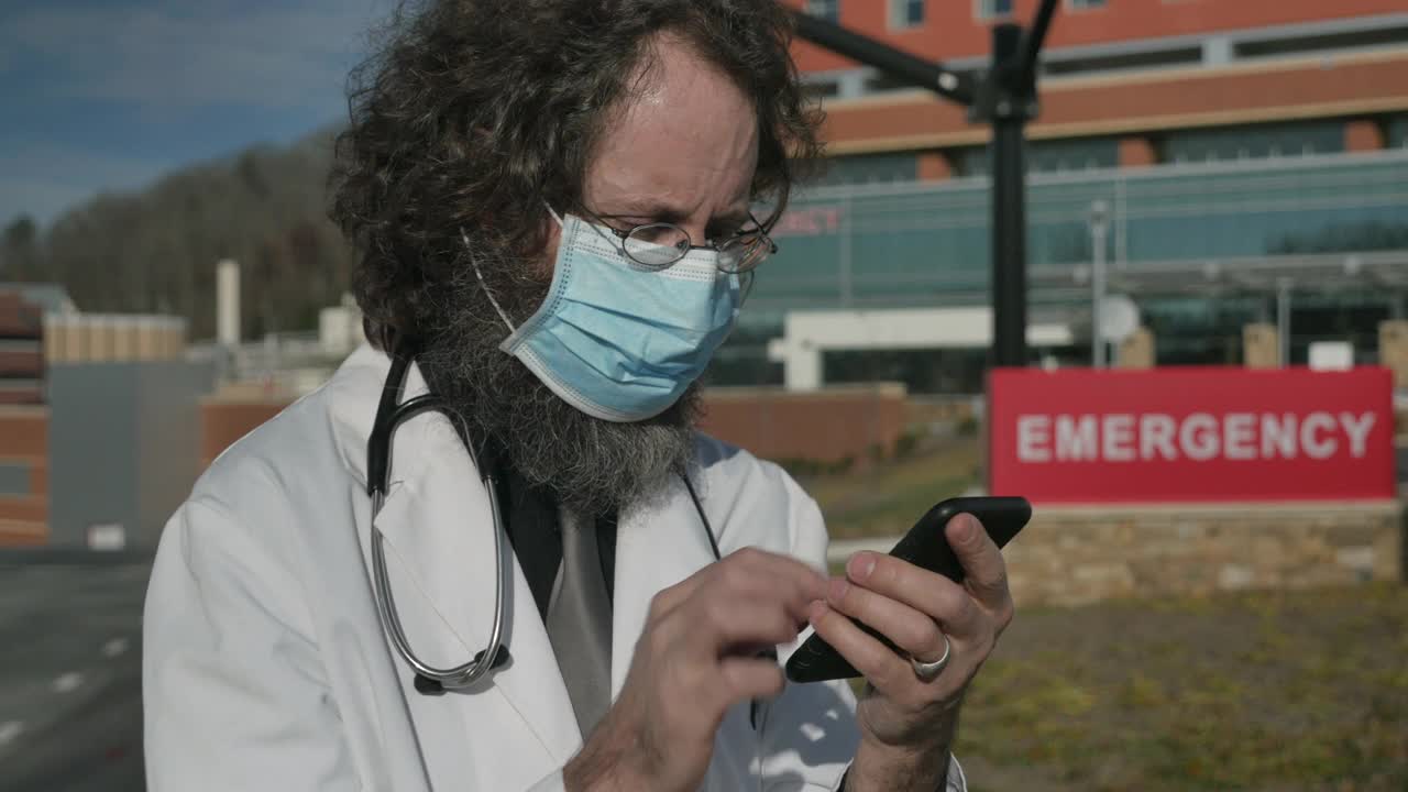 一个戴着口罩和他的手机交流的男医生的侧面视频下载