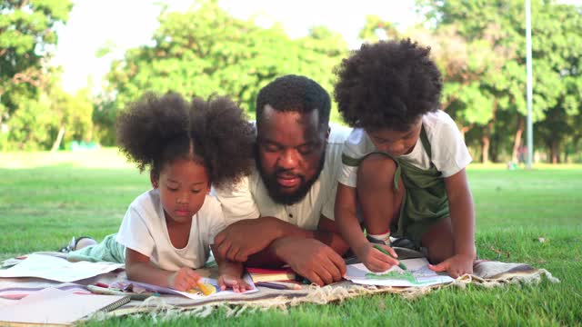 可爱的小黑人女孩，男孩和黑人父亲在阳光下享受野餐的一天。爸爸放下彩纸放松心情，和女儿、儿子一起创造创意。有两个孩子的家庭观念，生活简单。视频下载