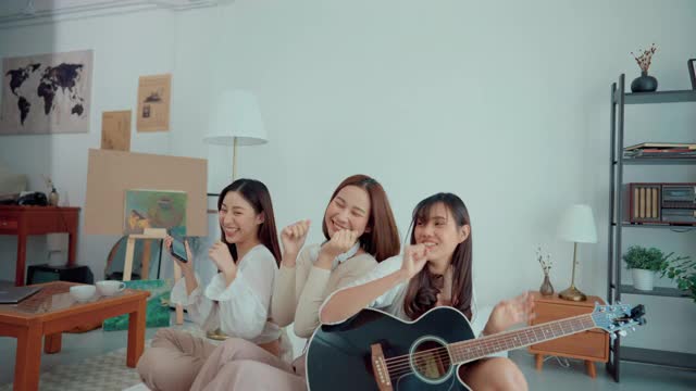 快乐的亚洲女人弹吉他和她的朋友唱歌跳舞在白色的公寓。视频素材