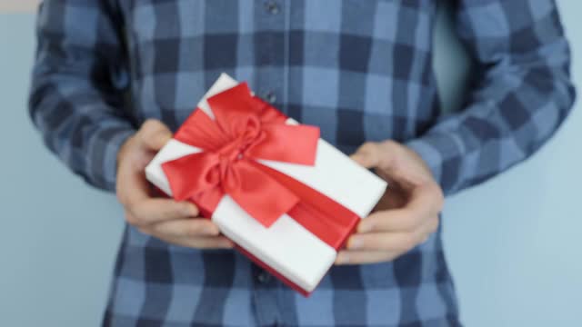 男子拿着并展示了系着红色蝴蝶结的白色礼品盒，走向镜头，特写。男性手中的情人节礼物。手正在赠送礼盒。视频下载