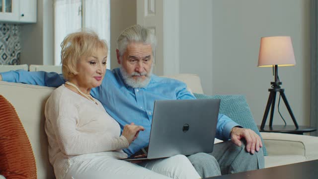 退休的老夫妇一起用笔记本电脑上网购物挑选特价商品，老人家的爷爷奶奶坐在家里的沙发上悠闲地看着电脑屏幕。冠状病毒隔离封锁。视频下载