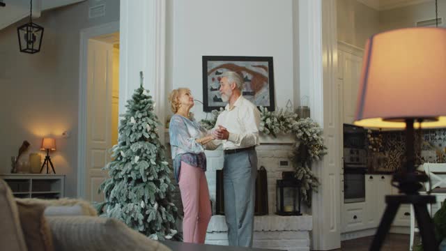 快乐的老女人和丈夫在装饰好的圣诞树前跳舞，模仿唱歌。有趣的退休人员在家里享受新年前夜的乐趣。视频下载