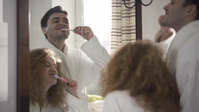 在浴室的镜子里，映照着早晨正在刷牙的英俊的白种人丈夫和漂亮的妻子。穿着白色浴袍的夫妇一家人。卫生及牙科护理视频素材